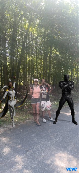 A photo of two people posing on a forest path with two digital avatars/edits—a robotic female character with a crescent blade on the left and a Black Panther character on the right—with a 'veve' watermark in the corner.