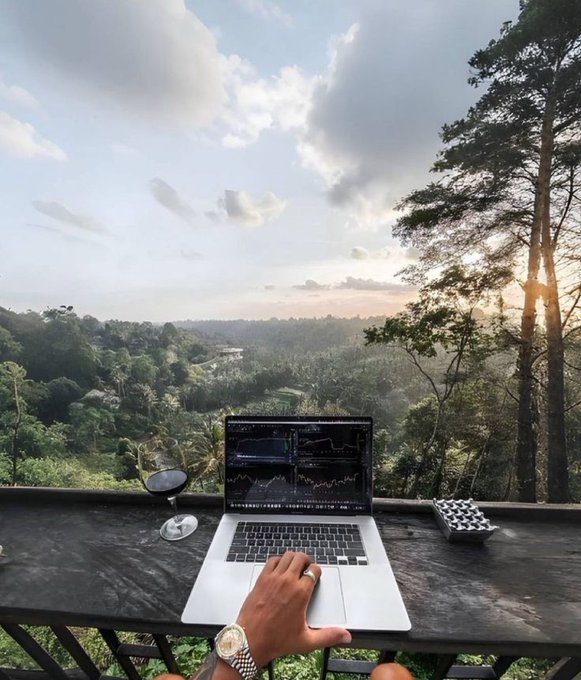 The image shows a person sitting at a table with their laptop open, likely working or browsing the internet. The table also has a wine glass and a cup of coffee placed next to the laptop. In the background, there are trees that provide a natural setting for this outdoor workspace. The photo is taken from an elevated position, possibly from a balcony or deck, giving a unique perspective on the scene.