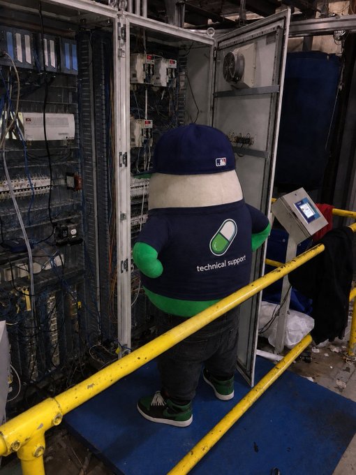 A person in a pill-shaped mascot costume wearing a "technical support" shirt and MLB cap stands before open industrial electrical panels inside the server room, humorously embodying the post's wordplay on "technical difficulties have been technically dealt with."