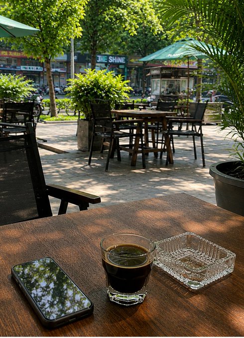 A wooden table at an outdoor cafe with a glass of dark liquid, an ashtray, and a smartphone, with trees and street shops visible in the background.