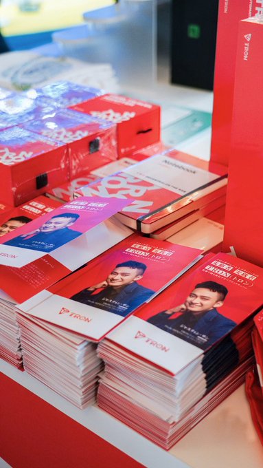 Stacks of red promotional brochures for 'TRON' featuring a man's portrait, displayed on a table at what appears to be a marketing event.