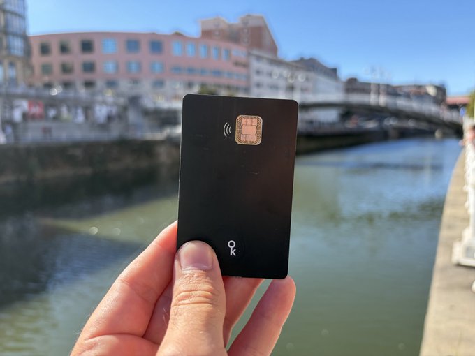 A hand holding a black card with an embedded chip and contactless symbol, featuring the "ok" logo, taken near a canal with a bridge and buildings in the background.