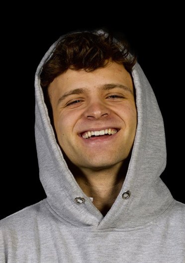 A young man with curly brown hair smiles brightly while wearing a gray hoodie with the hood up, set against a black background—likely representing a casual, relatable figure to accompany the news about Jack Maller's Bitcoin-yielding Strike platform.