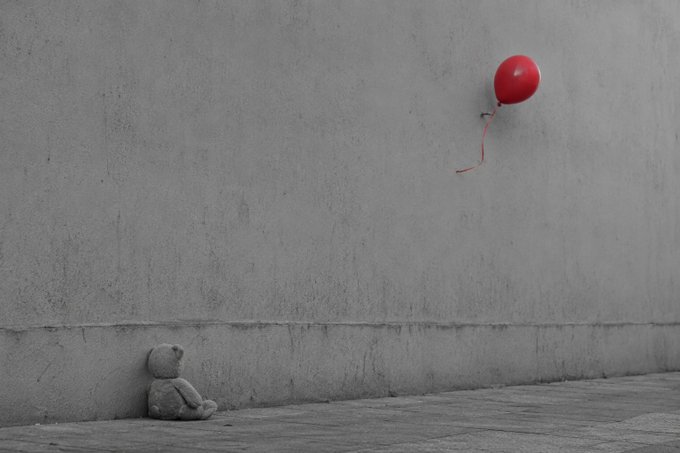 A black and white photograph showing a lone teddy bear sitting against a gray wall, looking up at a single red balloon floating away.