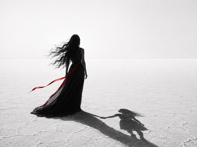 A minimalist black-and-white photograph of a woman in a long flowing black dress with a red ribbon, standing in a vast, cracked white salt flat with her long dark hair blowing in the wind, casting a dramatic shadow.