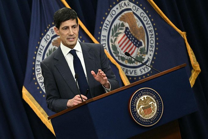 A man in a dark suit speaks at a podium bearing the seal of the Board of Governors of the Federal Reserve System, with the Federal Reserve seal flag displayed behind him.