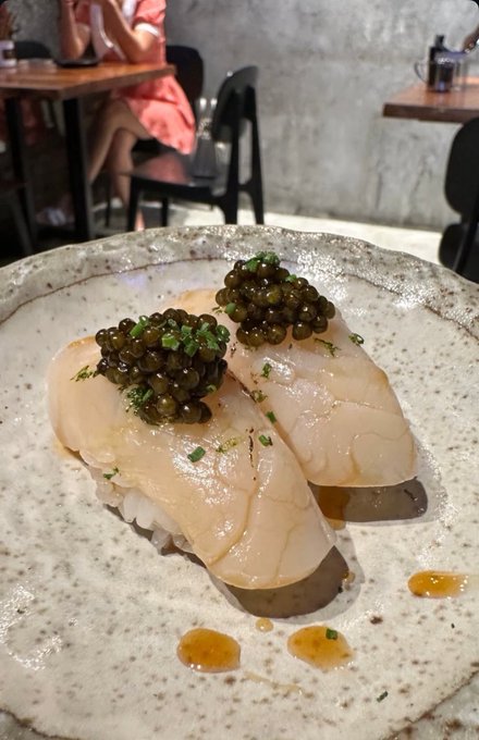 A close-up photograph shows two pieces of nigiri sushi topped with what appears to be caviar and microgreens, elegantly plated on a speckled ceramic dish with orange sauce drizzle.