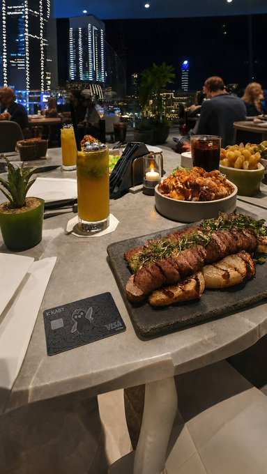 An evening dining scene at a rooftop or high-rise restaurant with a city skyline view, featuring a table spread with a grilled steak dish, fries, a salad, and an orange-colored drink in a tall glass, with a card reader on the table.