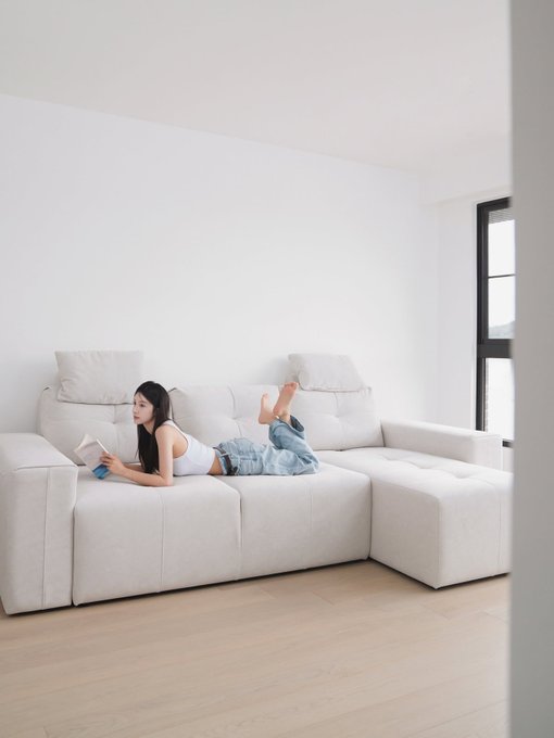 A young woman lies on a large white sectional sofa reading a book in a minimalist white room with light wood flooring.