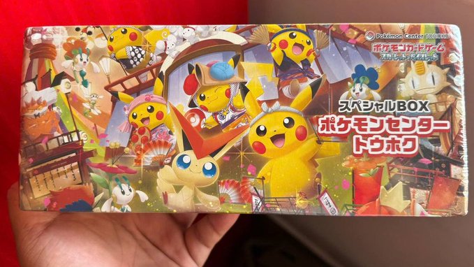 A hand holds a sealed Pokemon Center special box featuring Pikachu, Victini, and Shaymin with Japanese text reading 'スペシャルBOX ポケモンセンターバレンタイン' and 'ポケモンセンター ONLINE'.