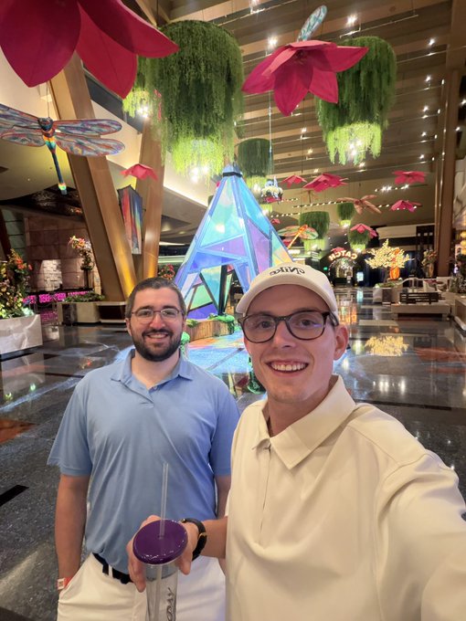 Two men pose for a selfie in a brightly decorated indoor venue with hanging pink flowers and greenery; one man wears a blue polo and holds a purple drink, while the other wears a white shirt and a 'PRIMERO' cap.