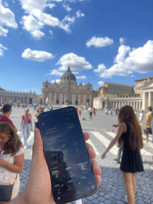 A person holds up a smartphone displaying a portfolio app showing 'Total Deposit $21,773.431' and 'Total Net Worth 4,308.691' in St. Peter's Square with the Vatican basilica visible in the background.