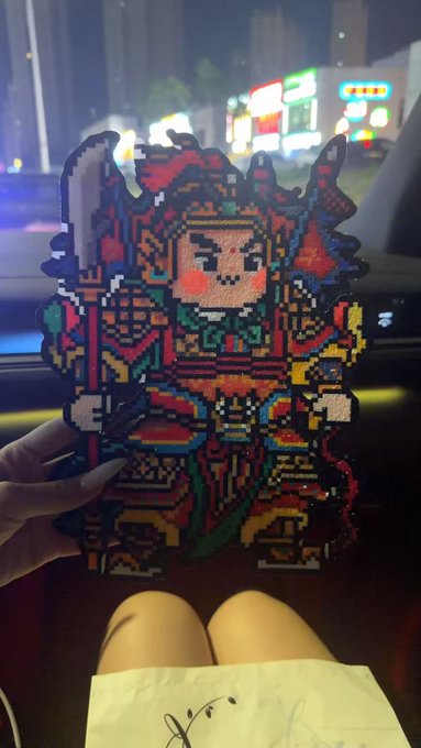 A person holds up a colorful pixel-art style leather craft depicting a fierce warrior character, taken from inside a vehicle at night. The intricate beadwork piece relates to the post's mention of "grinding a leather jacket," showcasing the handmade craft they completed while waiting for food during a late afternoon of crypto trading.