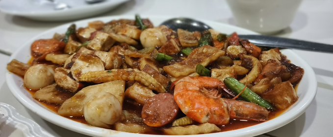 A plate of stir-fried food containing shrimp, squid, eggs, sausage, and vegetables in a savory sauce, served with a spoon on a white table.