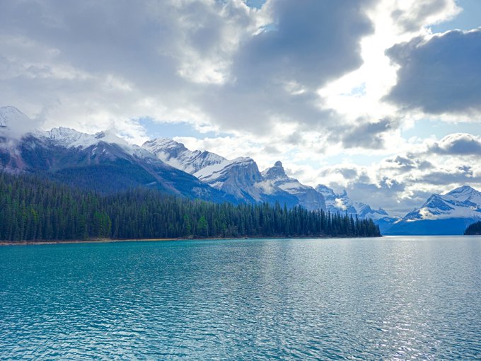 A scenic landscape photograph of a turquoise lake surrounded by evergreen forest with snow-capped mountain peaks under a partly cloudy sky.