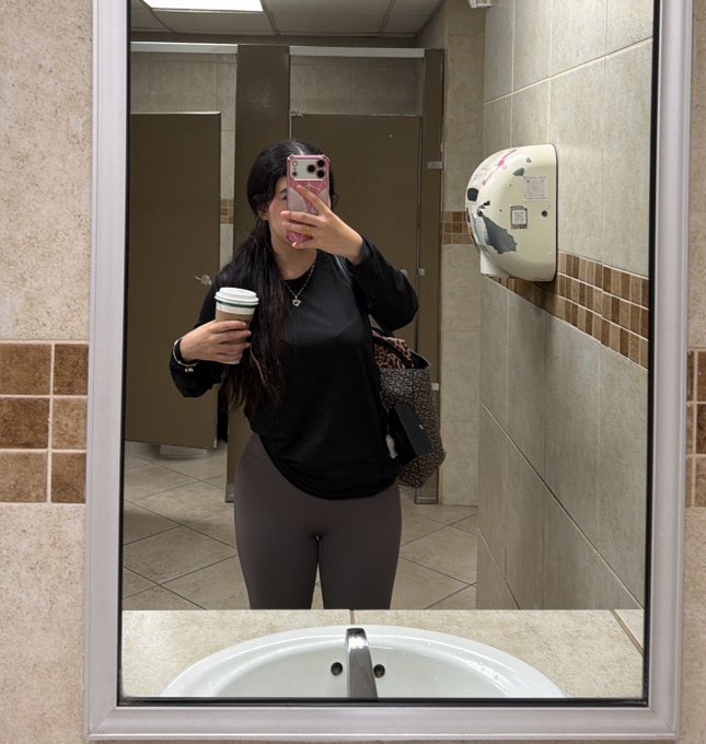 A woman takes a mirror selfie in a public restroom while holding a takeaway cup and carrying a patterned shoulder bag; a wall-mounted diaper-changing station is visible behind her.