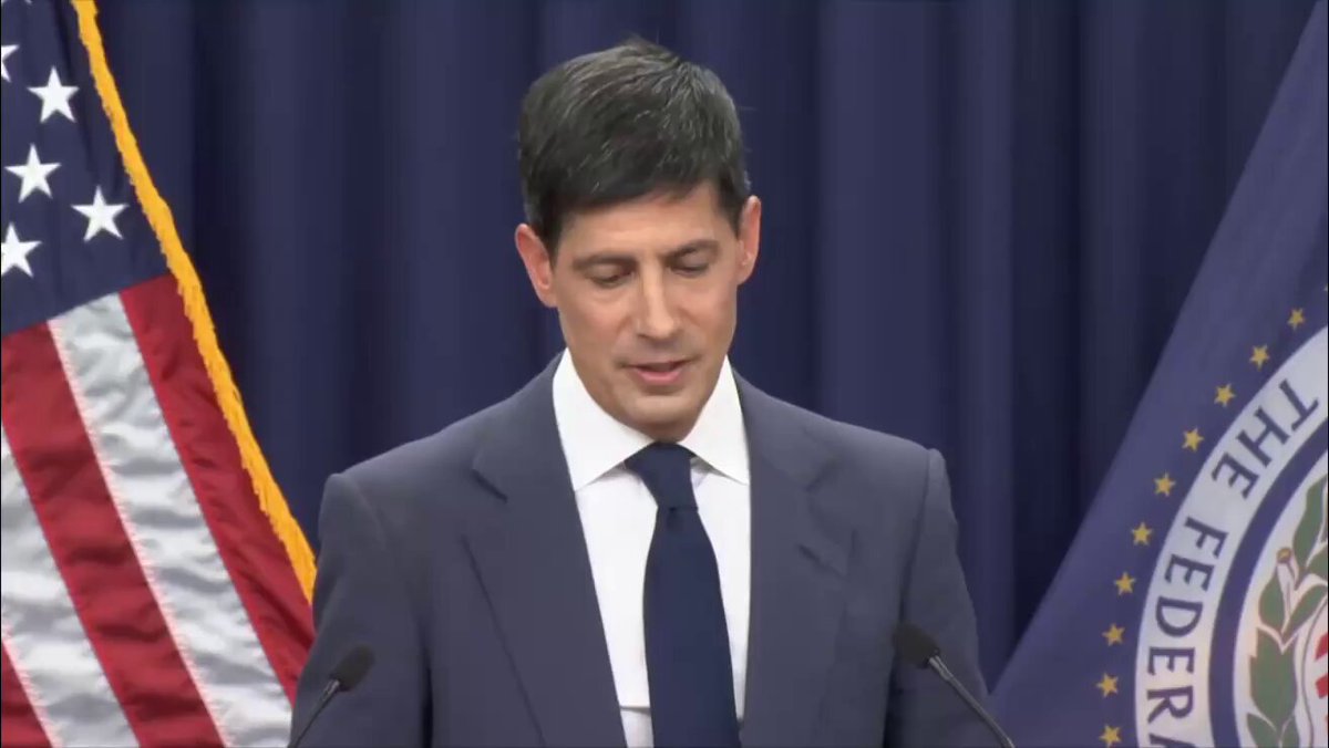 The image features a man dressed in a suit, standing at a podium with his hands clasped together as he speaks into a microphone. The backdrop of the scene includes an American flag and a blue curtain, suggesting that this event might be taking place within a government building or a similar official setting. The man's attire and the presence of the flag indicate that it could be related to a speech or a formal announcement made by a government official.