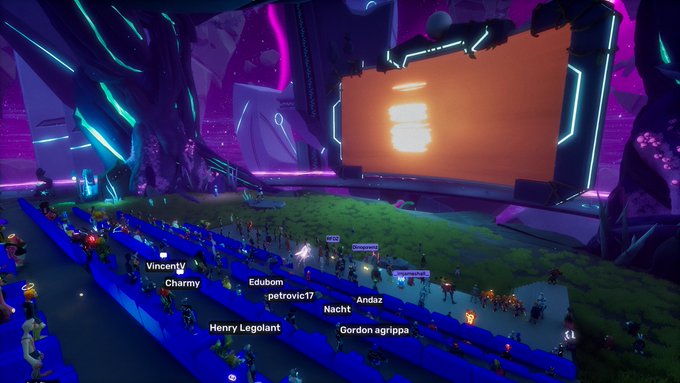 Avatars gather in a vibrant, neon-lit virtual theater to watch a premiere screening, with usernames like "VincentV," "Charmy," "Edubom," "petrovic17," "Andaz," "Nacht," "Henry Legoiant," and "Gordon agriopa" floating above the seated crowd in front of a glowing orange screen.
