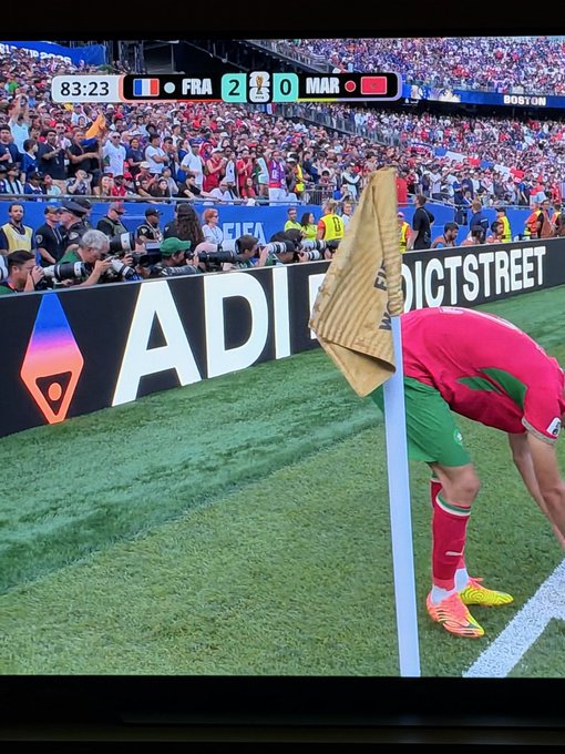 A player in a red jersey and green shorts bends over a corner flag on a soccer field, with a gloved goalkeeper's hand visible in the foreground. The scoreboard shows '83:23 FRA 2 - 0 MAR' with FIFA World Cup branding.