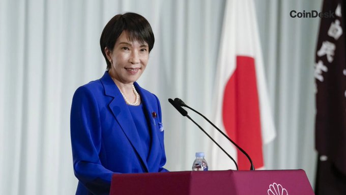 A Japanese official stands at a podium with the Japanese flag in the background, relating to Japan's plans for a national blockchain settlement system by the early 2030s. The CoinDesk logo is visible in the upper right corner.