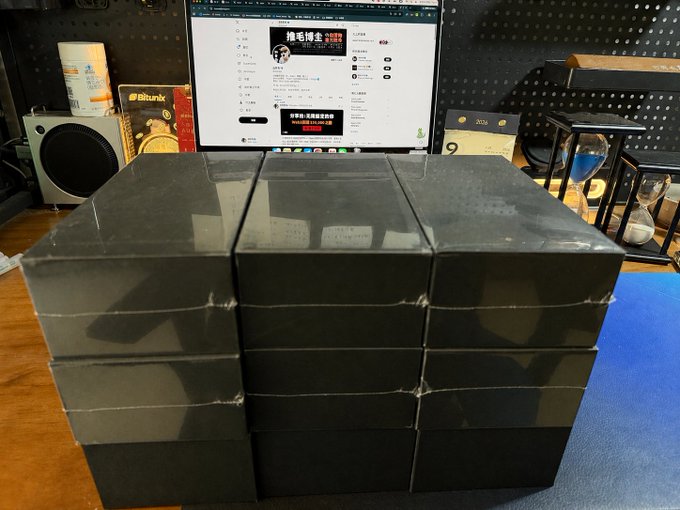 A stack of black boxes on a desk in front of a computer monitor displaying a webpage, with a speaker, books, and an hourglass visible in the background.