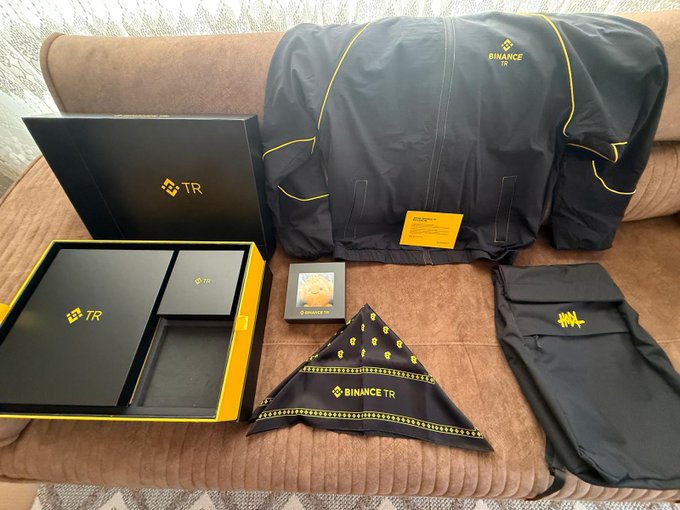 Binance merchandise including a black jacket with yellow accents, a bandana, and branded boxes are displayed on a couch.