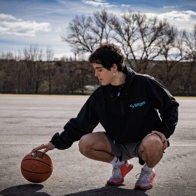 A young man crouches on an outdoor court, spinning an NBA-branded basketball while wearing a black "Bitget" windbreaker, channeling a relaxed pre-game energy that fits the vibe-focused caption.