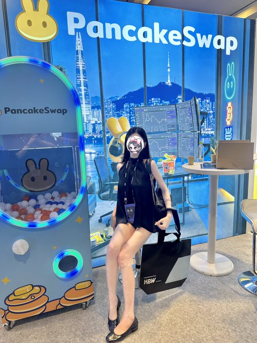 A woman in a black outfit and skull face mask poses next to a basketball game machine in front of a 'PancakeSwap' branded backdrop featuring a Seoul cityscape.