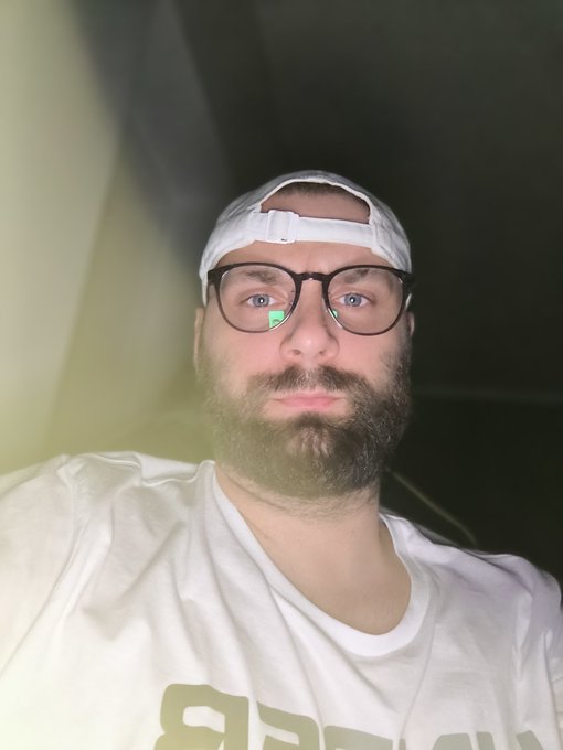In the image, there is a bearded man wearing glasses posing for a selfie with his face turned towards the camera. He's dressed casually in a white t-shirt that says "b" on it. The background of the photo appears to be dark, providing a contrasting backdrop to the subject and making him stand out. The post where this image was shared is titled "day 769 of posting selfie until $ZK hits $2 I expected it will be hard during bear but this is nuts 24hr price change +5% (current price 0.014)", which seems to have a connection with the photo's content or context, although we cannot determine its exact relevance from the image alone.