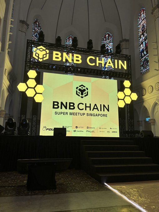 A stage is set up inside a building with stained glass and arched ceilings, featuring illuminated signage for "BNB CHAIN SUPER MEETUP SINGAPORE" alongside sponsor logos.