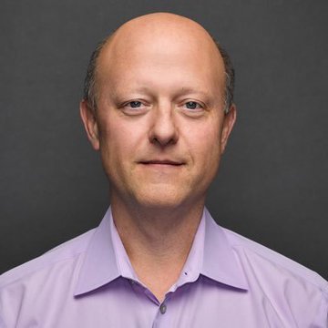 A professional headshot portrait of a bald man wearing a light purple dress shirt against a gray background.
