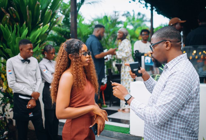A man in a checkered shirt interviews a woman with long curly hair in a rust-colored dress at the Arc Lagos event, while other attendees and staff in branded uniforms mingle in the lush, garden-like venue—capturing the "one-on-one conversations with Arc Builders" highlighted in the post.