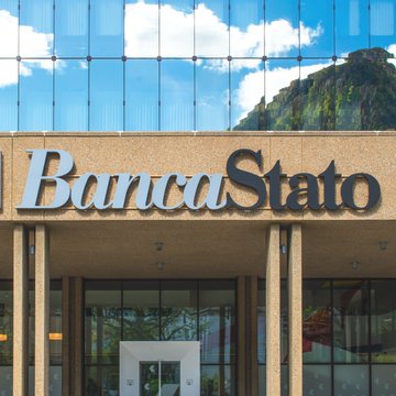 The image shows the entrance of BancaStato, a Swiss cantonal bank, with its name displayed in silver lettering against a stone facade beneath a modern glass building that reflects the surrounding mountains and sky. This visual ties directly to the post's announcement that BancaStato has become another traditional bank to embrace Bitcoin by launching regulated trading and custody services through Sygnum's infrastructure.