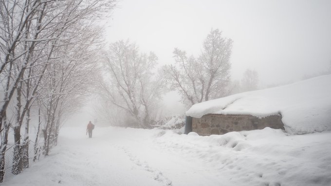 A misty, snow-covered winter landscape with a lone figure walking along a snowy path beside a stone structure and bare frost-covered trees.