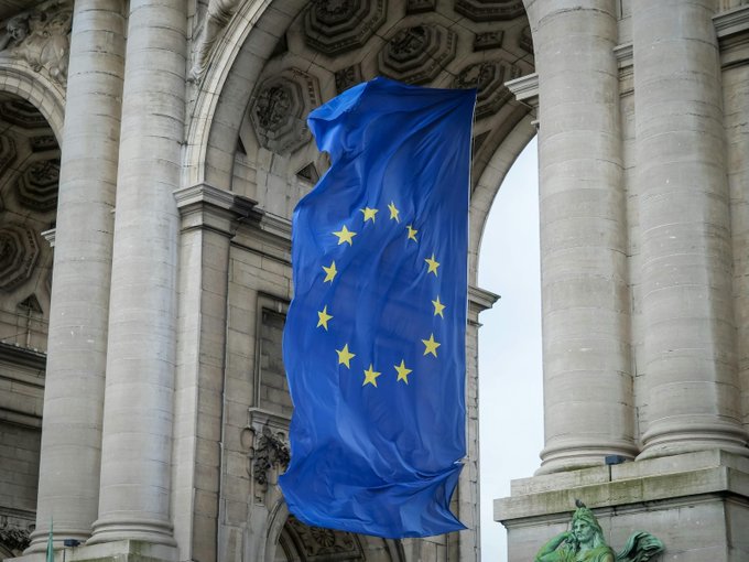 The European Union flag hangs prominently from an ornate stone archway, symbolizing the EU's regulatory body referenced in the post about ESMA's MiCA register expansion. The flag's twelve yellow stars on a blue field visually represent the growing European regulatory oversight, with 321 companies now listed.