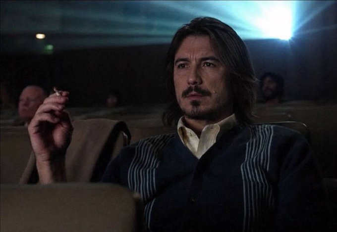 A man with shoulder-length dark hair, a mustache, and goatee, wearing a blue cardigan over a white shirt, sits in a dimly lit theater holding a cigarette.