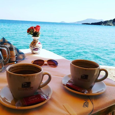 Two cappuccinos sit on a seaside table overlooking turquoise ocean waters, accompanied by sunglasses and a small vase of flowers, with the "Sheraton.com" logo visible on the coffee cups—perfectly capturing the relaxed, "good morning" vibe of the post.