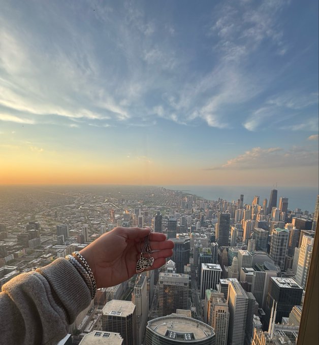 A hand holding a silver pendant extends outward against a panoramic sunset view of a sprawling city skyline, likely Chicago.