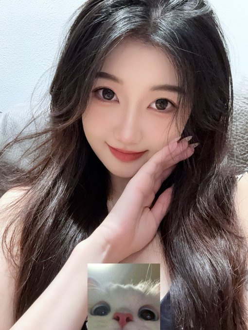 A young woman with long black hair poses with her hand near her face, smiling sweetly at the camera, while a small inset image of a wide-eyed white cat with a surprised expression appears at the bottom—humorously conveying the "shock" of being reminded that a promised gift still hasn't been delivered.