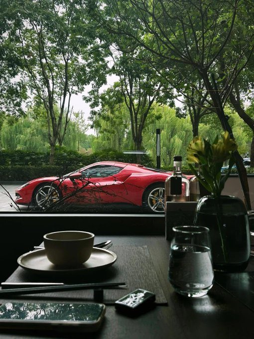 The image shows a dining table with various items, including a bowl of food, a cell phone, a glass vase with flowers, and a car outside. The caption reads "Be honest. What do you need most right now? 100K XRP 2 $BTC Or...". This suggests that the person in front of the image might be contemplating their financial situation or considering investing in cryptocurrency like Bitcoin.