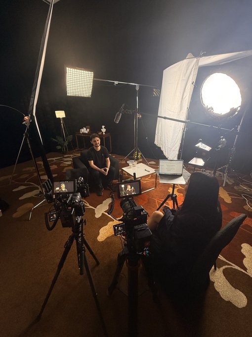Behind-the-scenes view of a professional photo/video studio setup with multiple cameras on tripods, large softbox lights, and a subject seated in the center being filmed by a videographer.