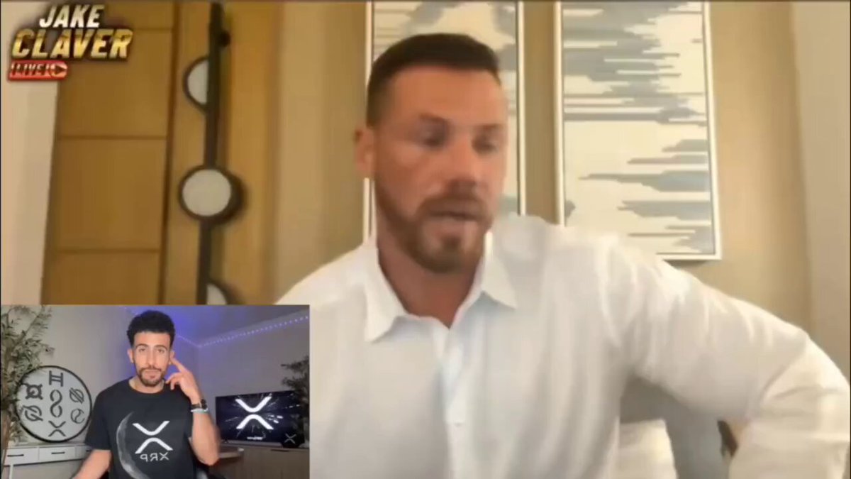 The image features a man wearing a white shirt sitting at a desk with a computer monitor behind him. The background of the image shows a wall with a painting, and there are two clocks visible - one near the top left corner and another on the right side of the image. The main focus is on the man in the white shirt who appears to be talking or giving a speech at a conference.