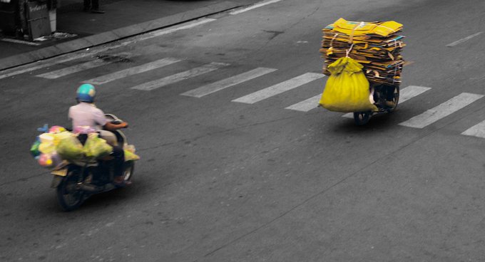 Two motorcyclists ride across a crosswalk on a city street; one carries colorful stuffed toys while the other transports large stacks of yellow packages.