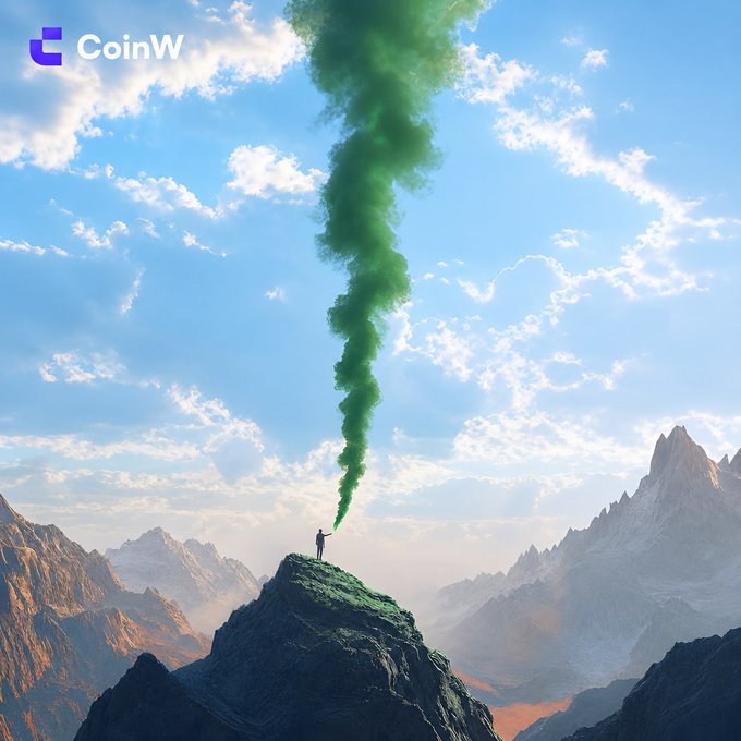 A lone figure stands atop a mountain peak releasing a plume of green smoke into a vast blue sky, with the "CoinW" logo displayed in the upper left corner. The triumphant summit moment visualizes the upbeat "GM" (good morning) spirit, symbolizing reaching new heights in the crypto market.