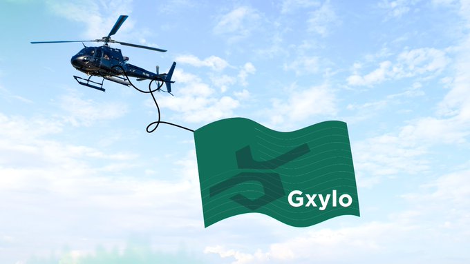 A blue helicopter flies through a partly cloudy sky, towing a large green flag that displays "Gxylo" alongside a stylized "5" logo, whimsically fulfilling the wish for a creative promotional display.