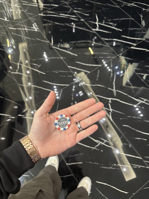 A person's hand holding a casino chip with "5,000" printed on it, set against a shiny black marble floor.