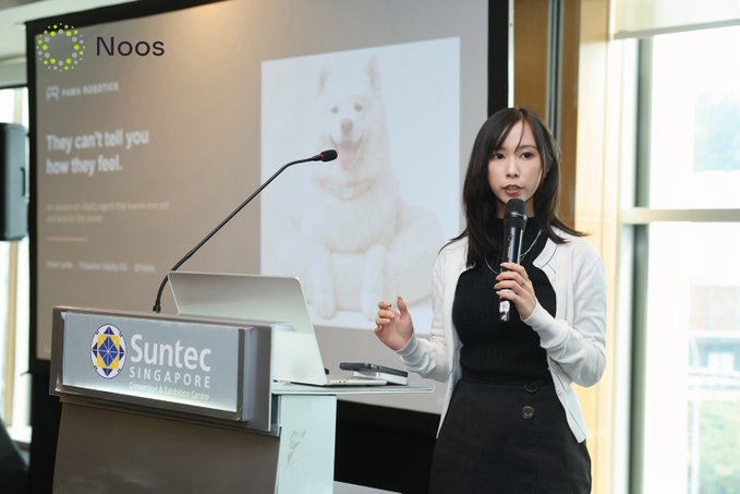 A woman in a black dress and white cardigan speaks into a microphone at a Suntec Singapore podium, with a screen behind her displaying the Noos logo, a picture of a Shiba Inu dog, and the text 'They can't tell you how they feel.'
