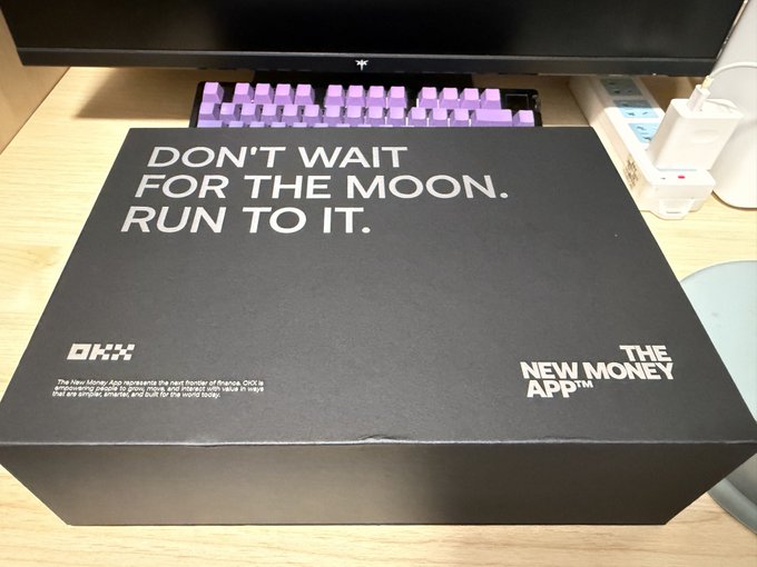 A black box on a desk with the text "DON'T WAIT FOR THE MOON. RUN TO IT." alongside the "OKX" logo and "THE NEW MONEY APP™" branding, with a pink keyboard and monitor in the background.