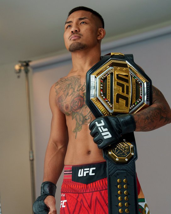 Joshua Van poses confidently with the UFC bantamweight championship belt over his shoulder, wearing UFC fight gloves and red-and-black patterned shorts, with visible tattoos on his chest and arm as he looks upward ahead of UFC 331.