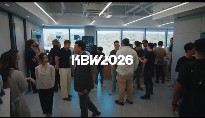 A group of people mingling in a modern office space with large windows, overlaid with white text reading 'KBW2026'.